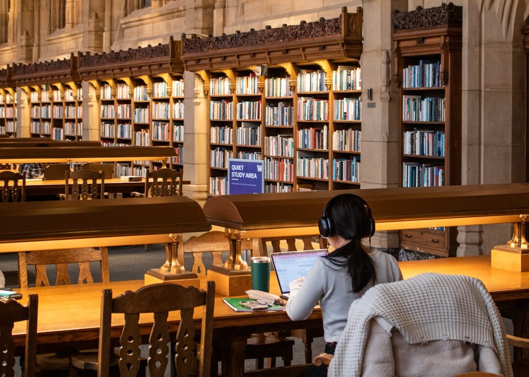 a young woman sitting in a library, surrounded by books, studying on her laptop